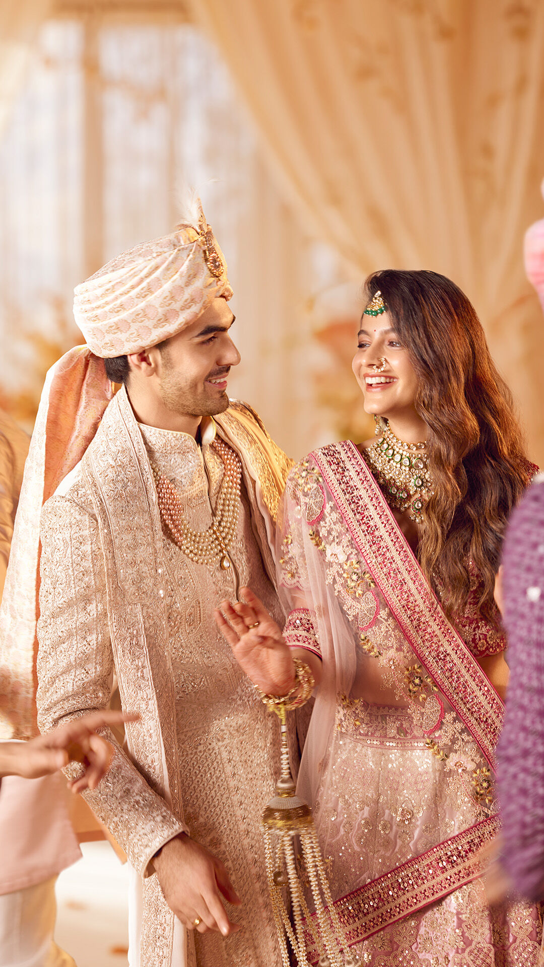 Smiling bride and groom in pastel pink and gold traditional wedding attire, looking at each other joyfully during an indoor wedding ceremony.